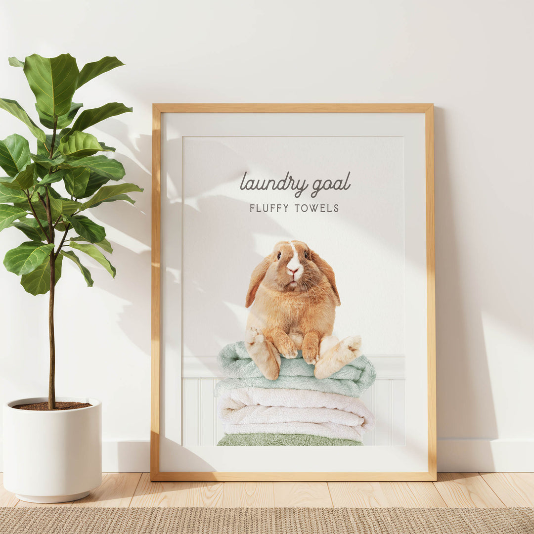 A framed image of a fluffy towel with a rabbit sitting on top of it, placed on a wooden floor next to a potted plant.