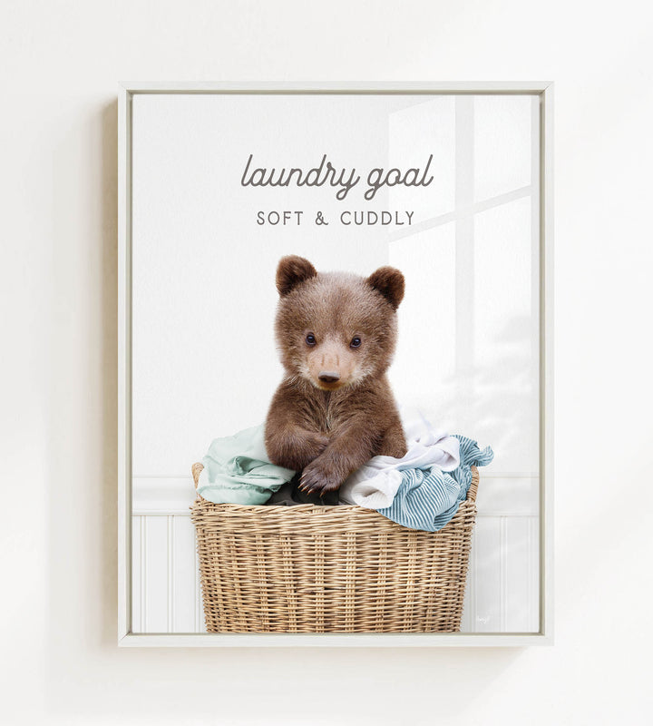 A cute brown bear sitting in a wicker laundry basket filled with clothes, with the text "laundry goal" and "soft & cuddly" written above it.
