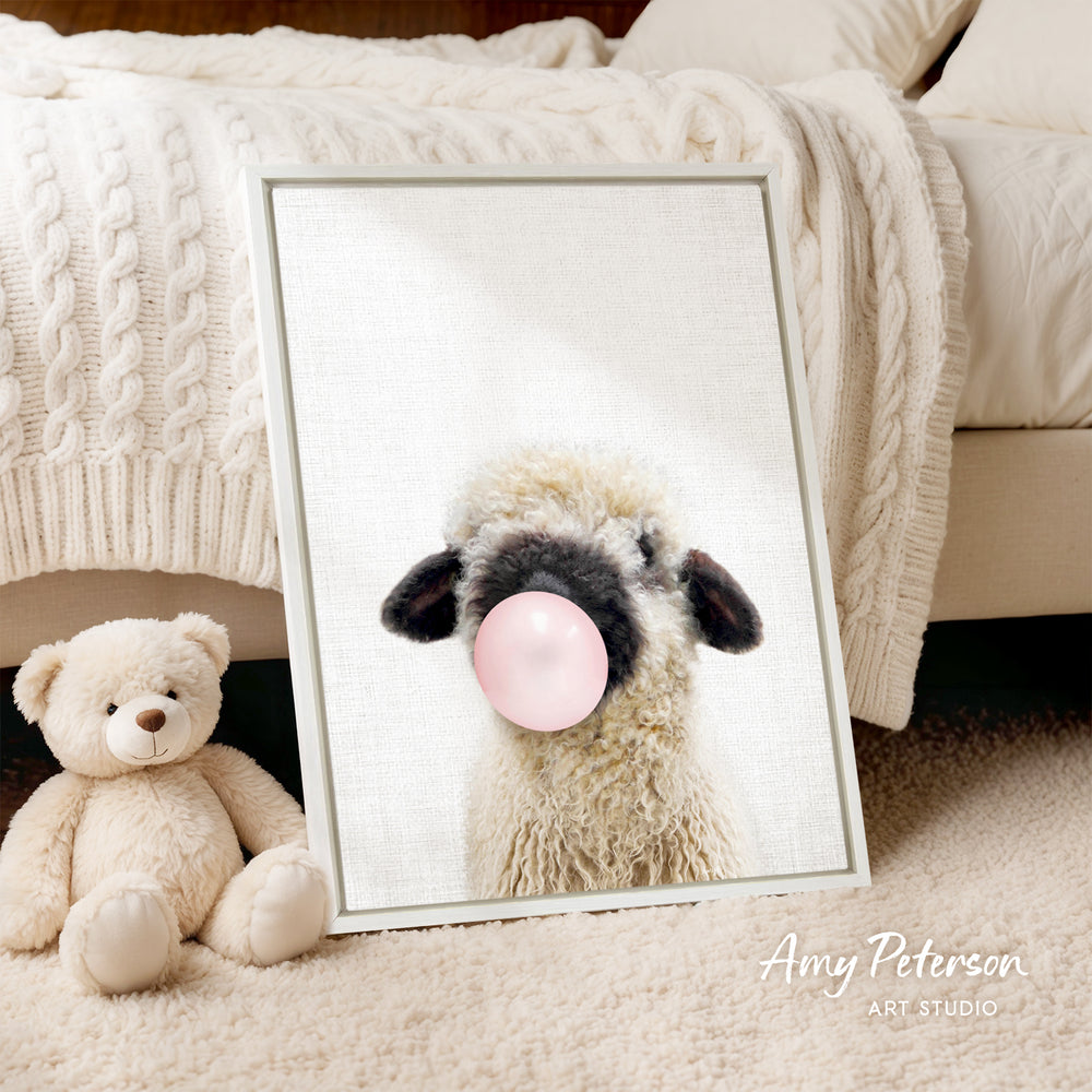 A framed image of a sheep blowing a bubble gum bubble is displayed on a bed, accompanied by a teddy bear.