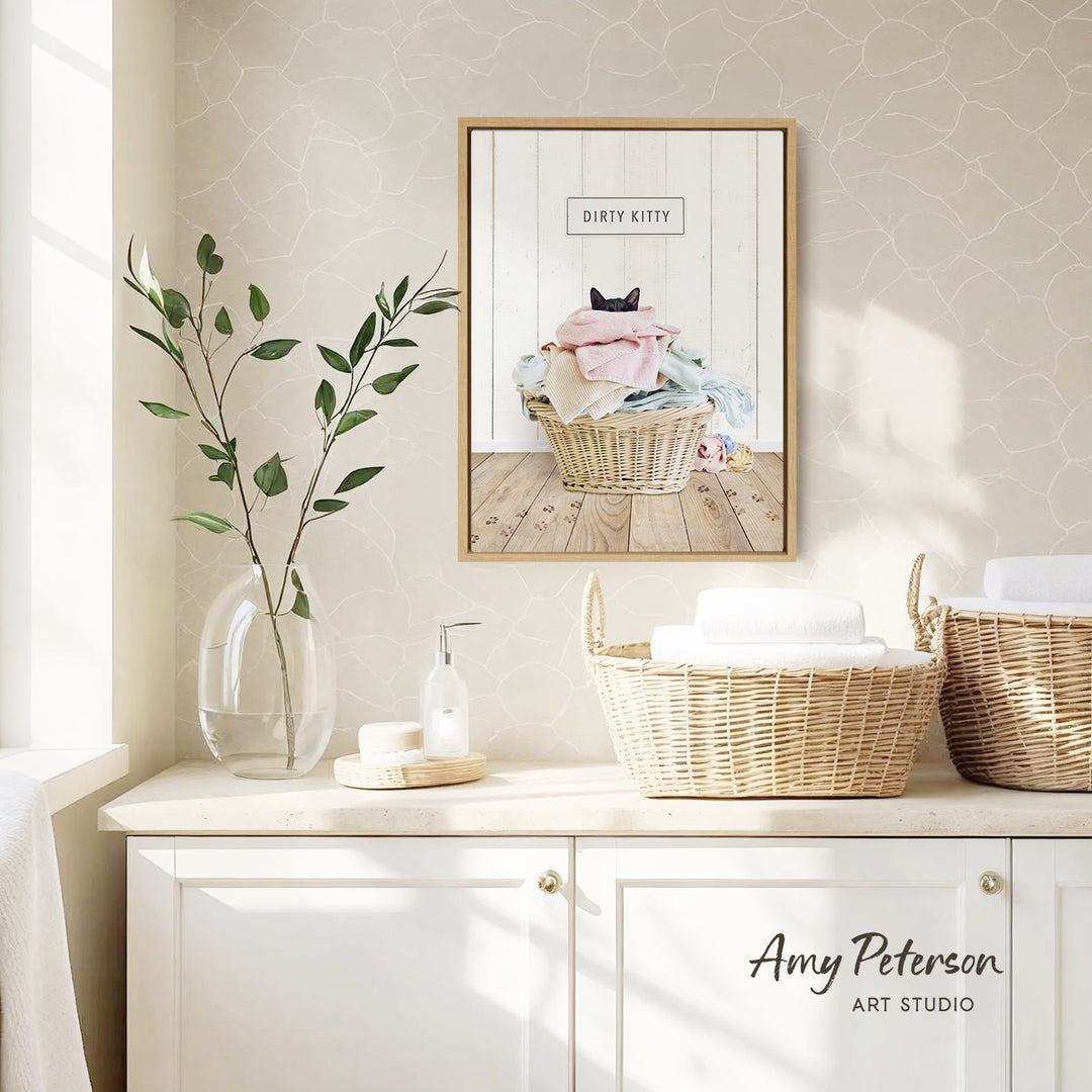 a bathroom setting with a framed print of a basket with a cat inside, a vase with green leaves, and baskets with towels.