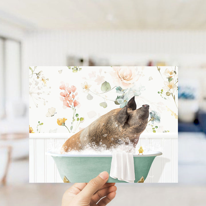 A hand holding a framed print featuring a pig sitting in a bathtub filled with bubbles, surrounded by a floral pattern.