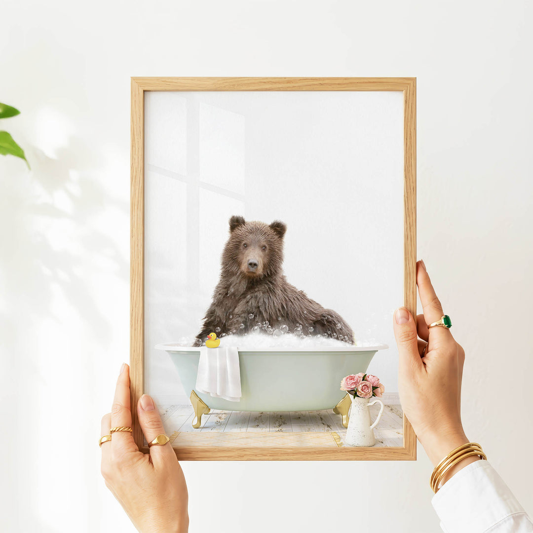 A person's hands are holding a picture frame with a framed image of a bear sitting in a bathtub, surrounded by flowers and a yellow rubber duck.
