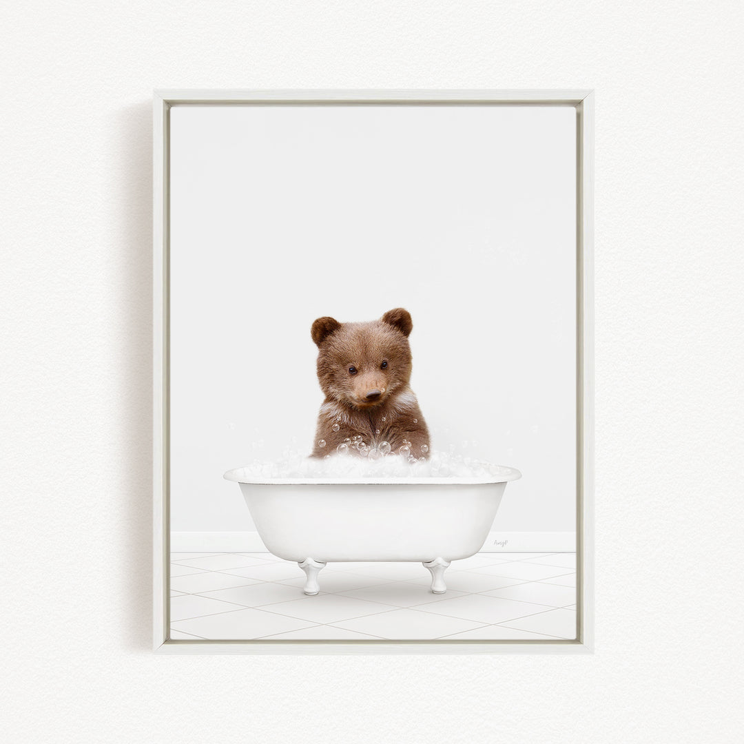 A cute brown bear sitting in a white bathtub filled with bubbles, looking directly at the camera.