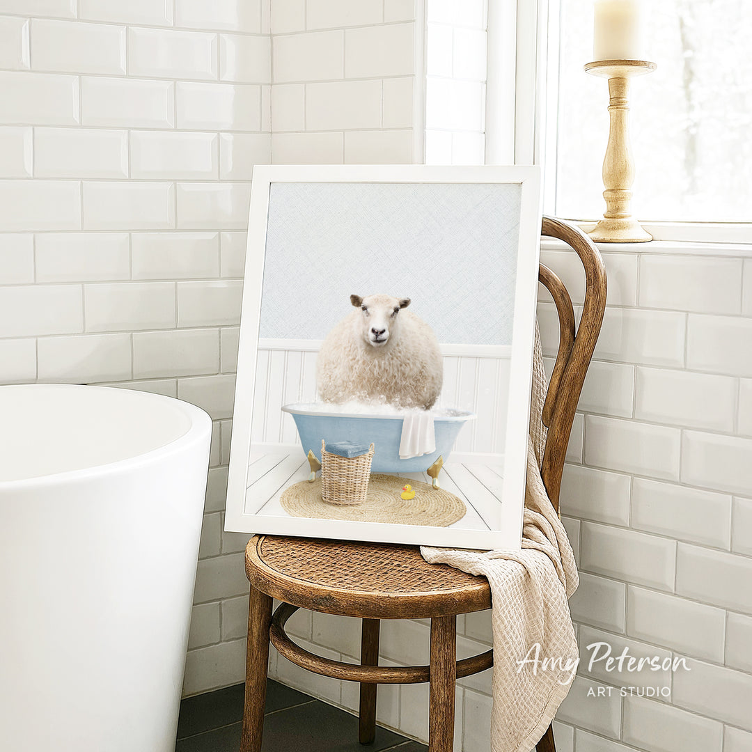 A framed image of a sheep sitting in a bathtub, surrounded by a basket of bath toys, is displayed on a wooden chair in a bathroom setting.