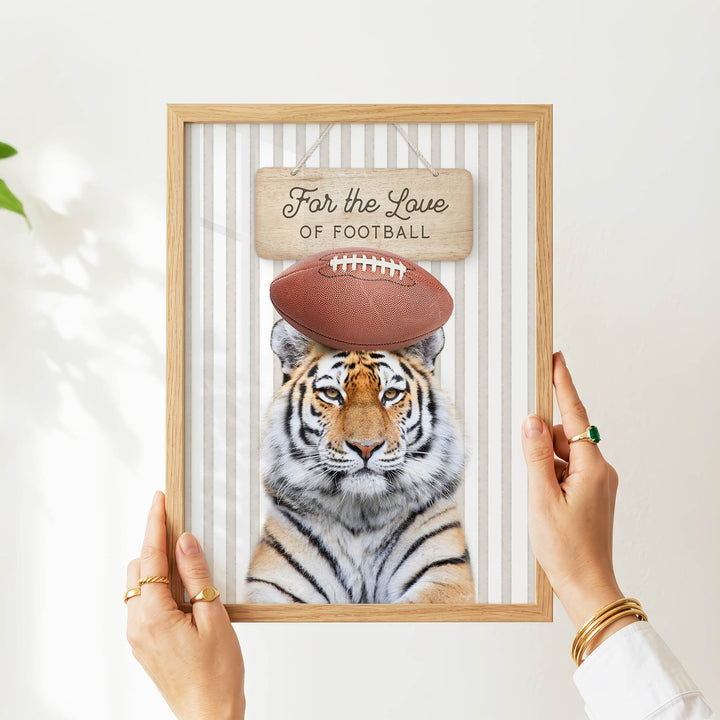 A person's hands are holding a framed picture of a tiger wearing a football helmet, with a sign above the tiger that says "For the Love of Football".