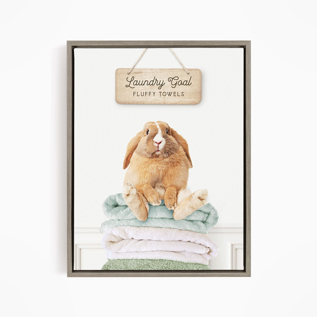 A wooden sign with the text "Laundry Goal Fluffy Towels" hangs above a stack of folded towels, with a rabbit sitting on top of the towels.