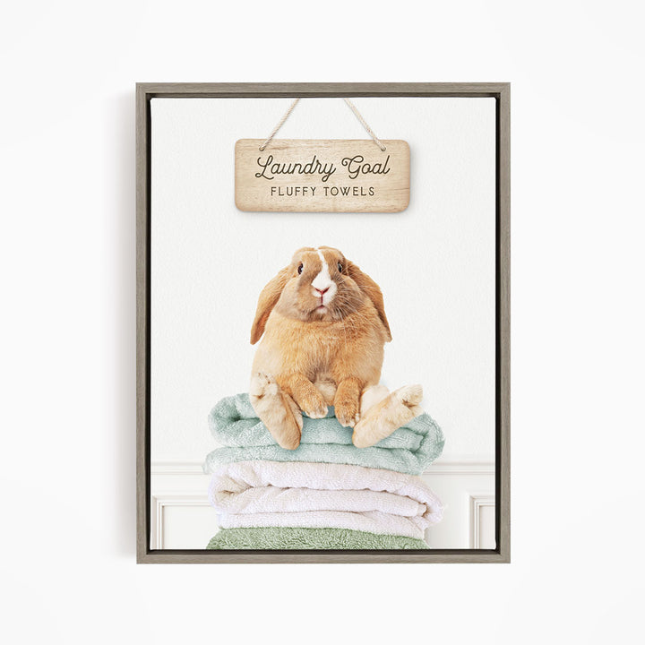 A wooden sign with the text "Laundry Goal Fluffy Towels" hangs above a stack of folded towels, with a rabbit sitting on top of the towels.