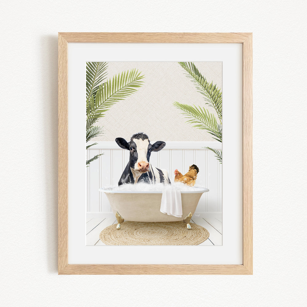 A framed image depicting a cow sitting in a bathtub, surrounded by palm leaves, with a chicken nearby.