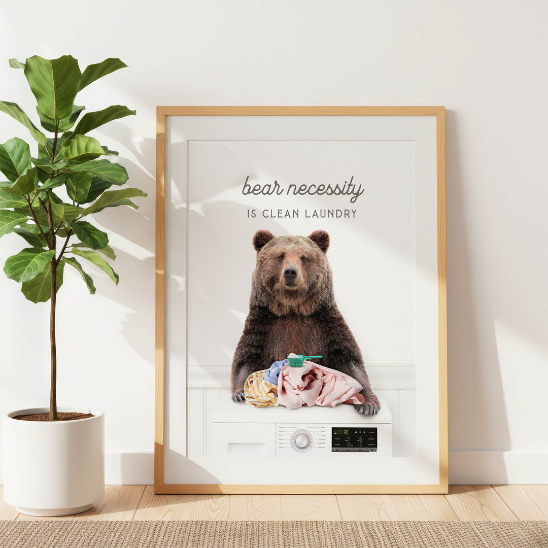 A framed poster with a brown bear sitting on a washing machine, with the text "bear necessity is clean laundry" displayed above the bear.