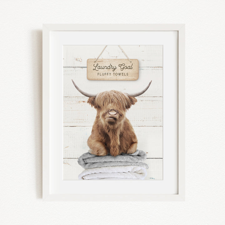 A framed image of a brown cow with long horns sitting on a stack of towels, with a sign above it that says "Laundry Coal Fluffy Towels".