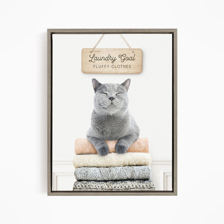 A gray cat sitting on a stack of folded clothes, with a wooden sign above that says "Laundry Goal Fluffy Clothes".