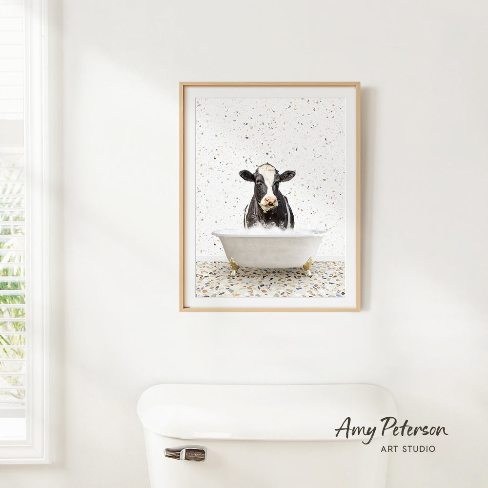 A framed print of a cow sitting in a bathtub is hanging on a white wall.