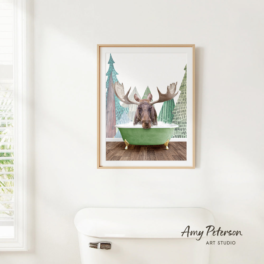 A framed print of a moose sitting in a green bathtub, surrounded by pine trees, is hanging on a white wall.