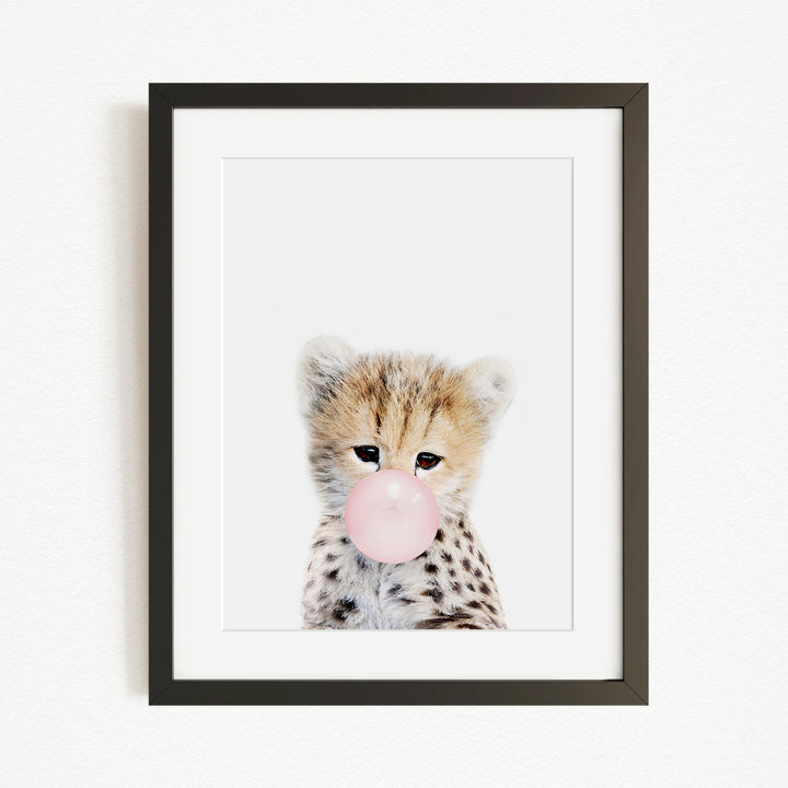 A young cheetah with spotted fur blowing a bubble with a pink bubble gum bubble.