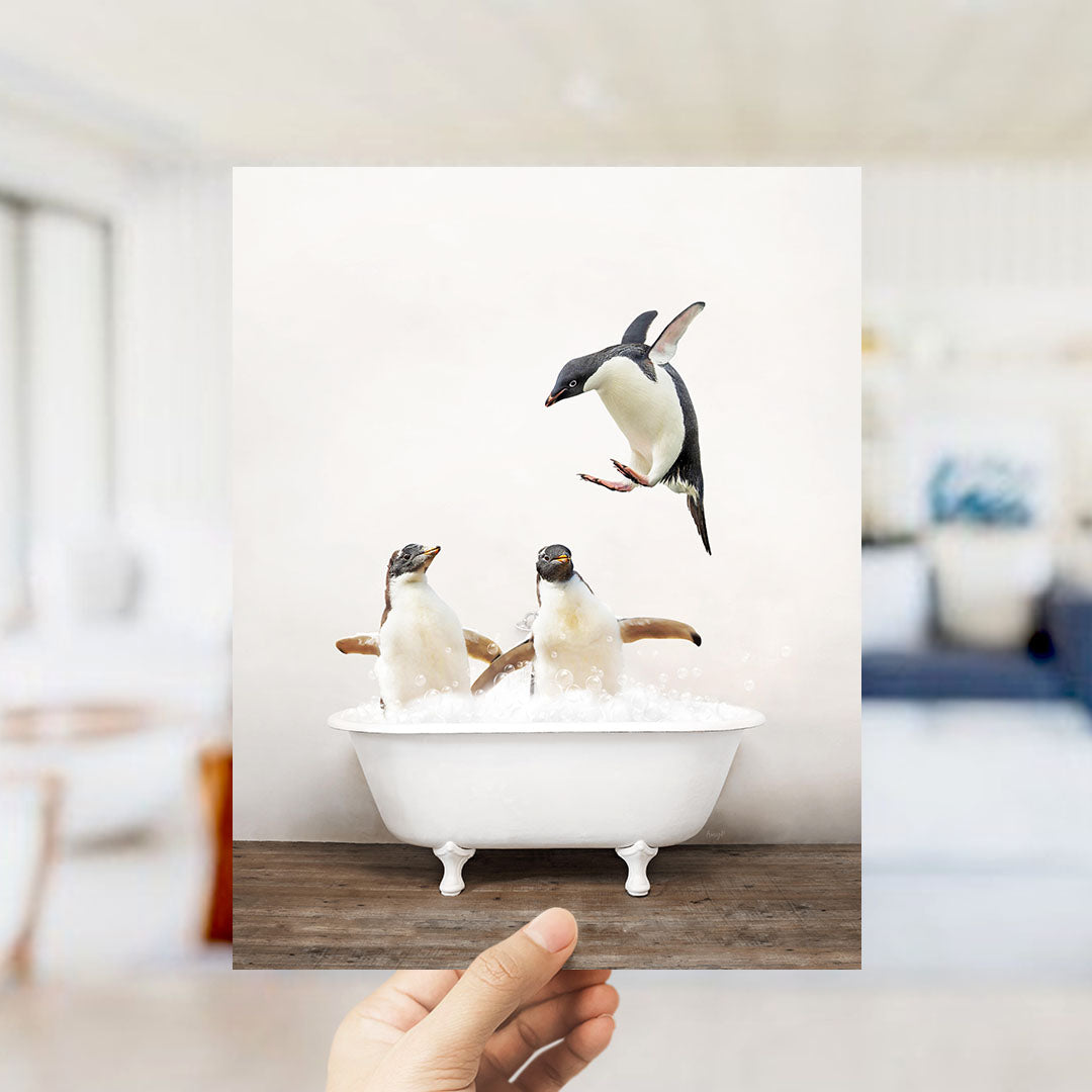 A hand holds up a framed photograph of two penguins sitting in a bathtub, with one penguin flying towards the other.