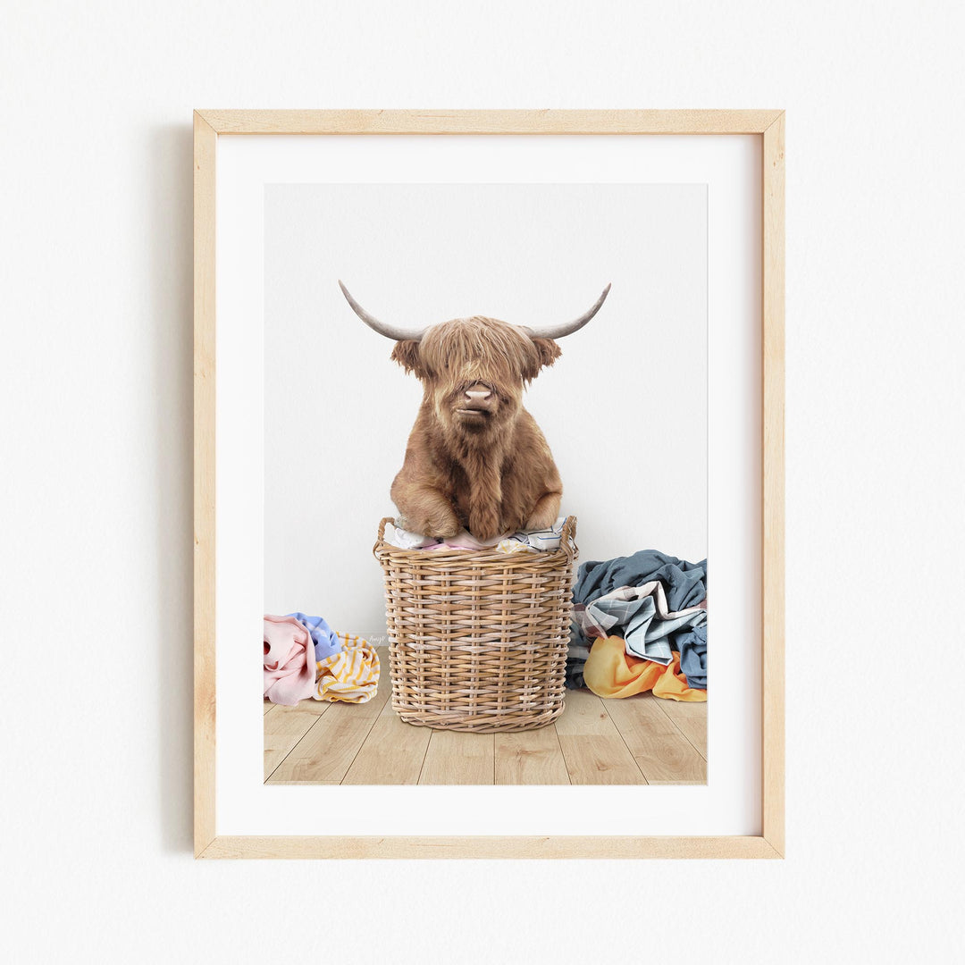 A Highland cow with long horns is sitting in a wicker basket filled with clothes, creating a whimsical and playful scene.