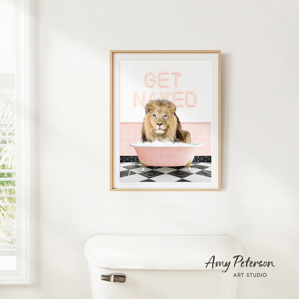A framed print of a lion sitting in a pink bathtub with the text "GET NAKED" on it, hanging on a white wall.