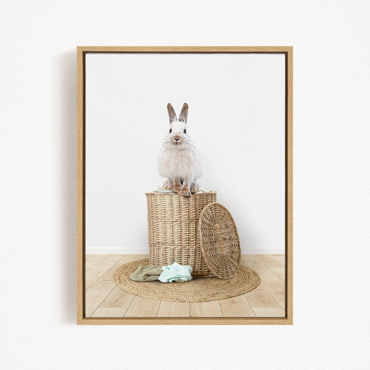 A white rabbit sits atop a woven basket on a wooden floor, with a green blanket and a basket lid nearby.