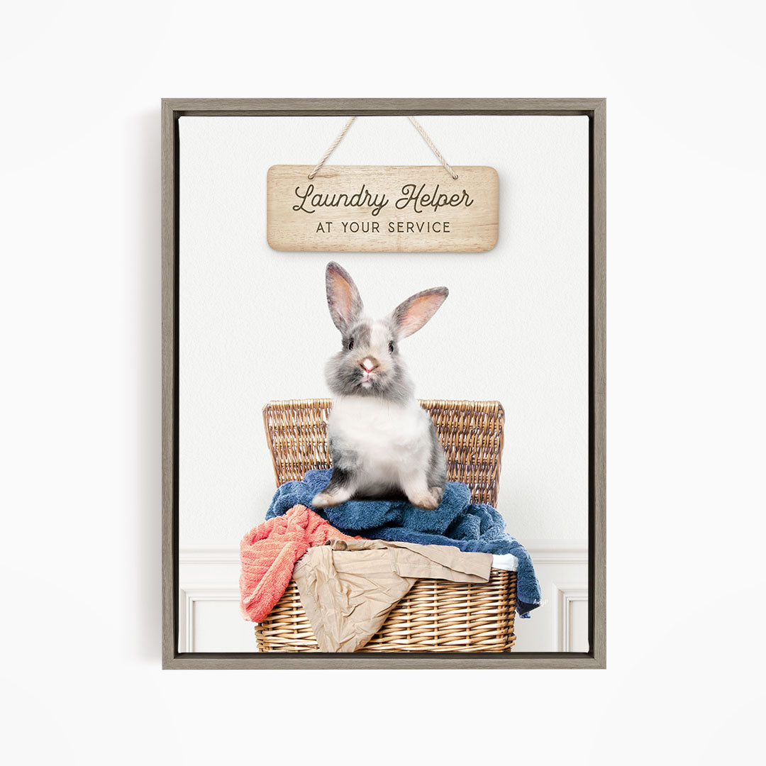 A gray and white rabbit sitting in a wicker laundry basket, with a wooden sign above that reads "Laundry Helper at your service".