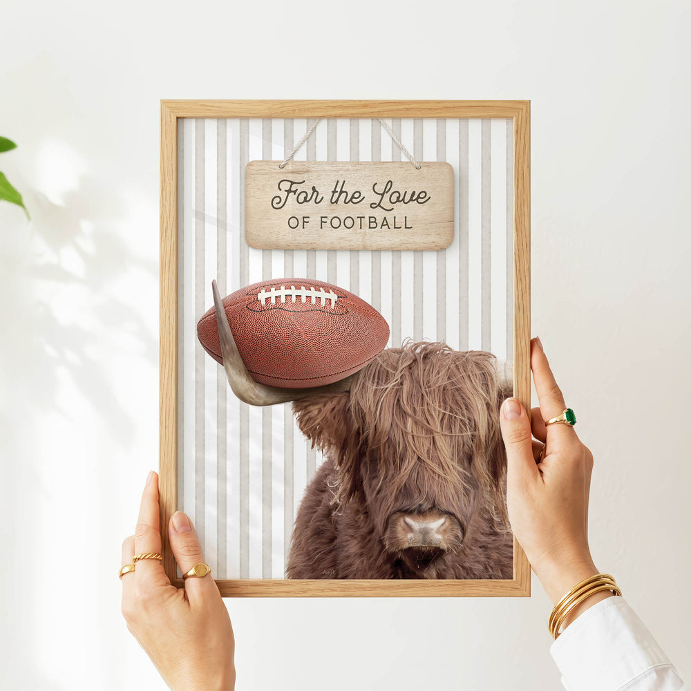 A person's hands are holding a framed picture of a Highland cow with a football on its head, accompanied by a wooden sign that reads "Far the Love of Football".