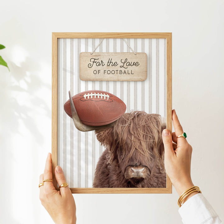 A person's hands are holding a framed picture of a Highland cow with a football on its head, accompanied by a wooden sign that reads "Far the Love of Football".