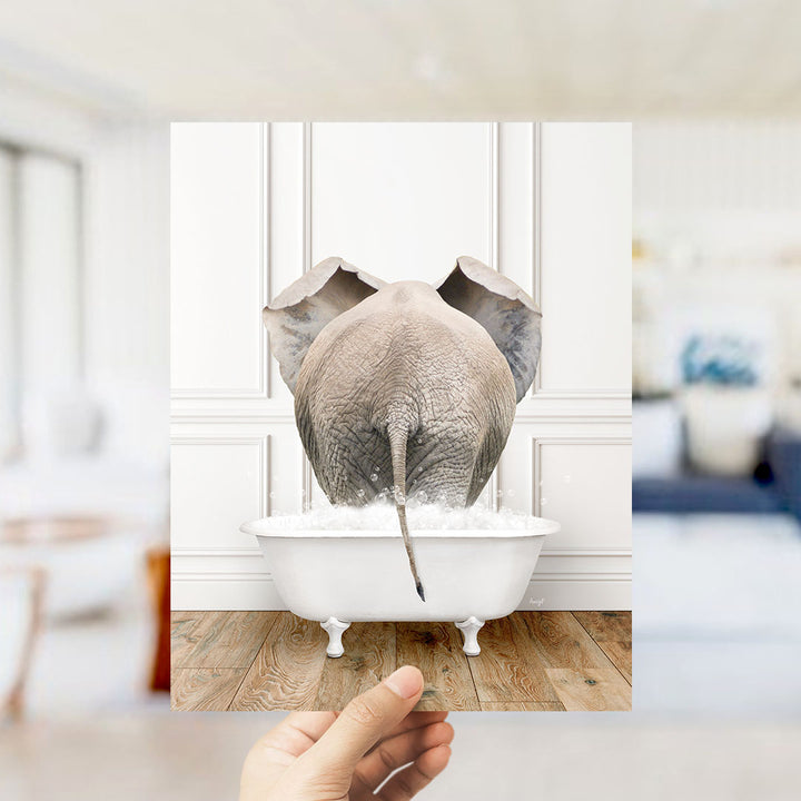A hand holds up a framed photograph of an elephant standing in a bathtub filled with water, creating a whimsical and playful scene.