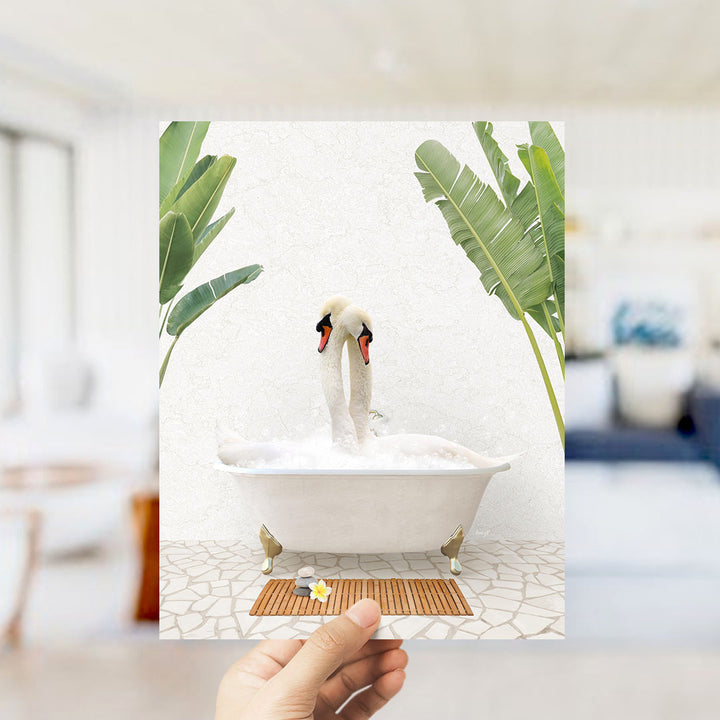 A hand holds up a framed photograph of a pair of swans sitting in a bathtub, surrounded by tropical plants.