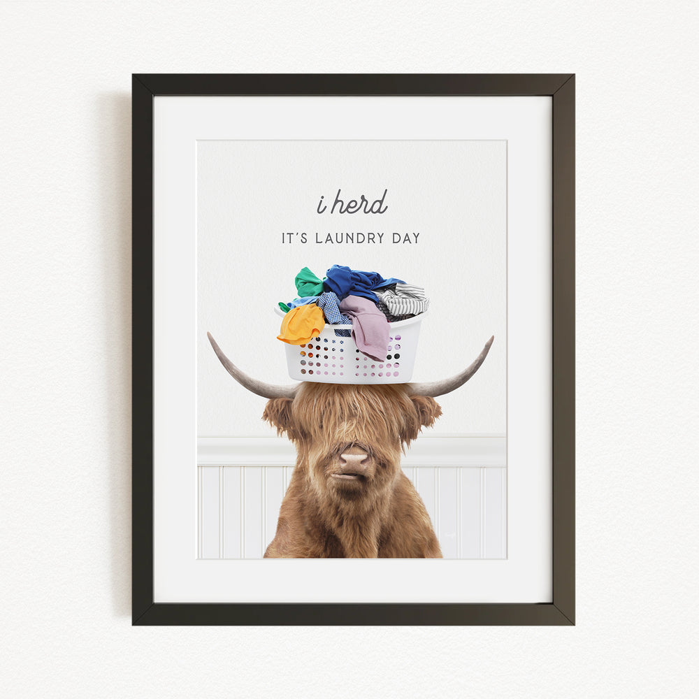 A framed image of a Highland cow with a basket of laundry on its head, accompanied by the text "It's laundry day"