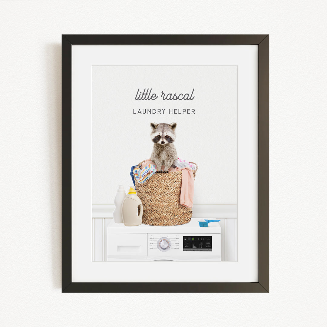 A raccoon sitting in a laundry basket filled with clothes and detergent, with a washing machine nearby.