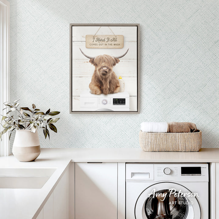 a framed poster of a Highland cow with horns, hanging on a wall above a laundry basket and a washing machine in a room with a white countertop.