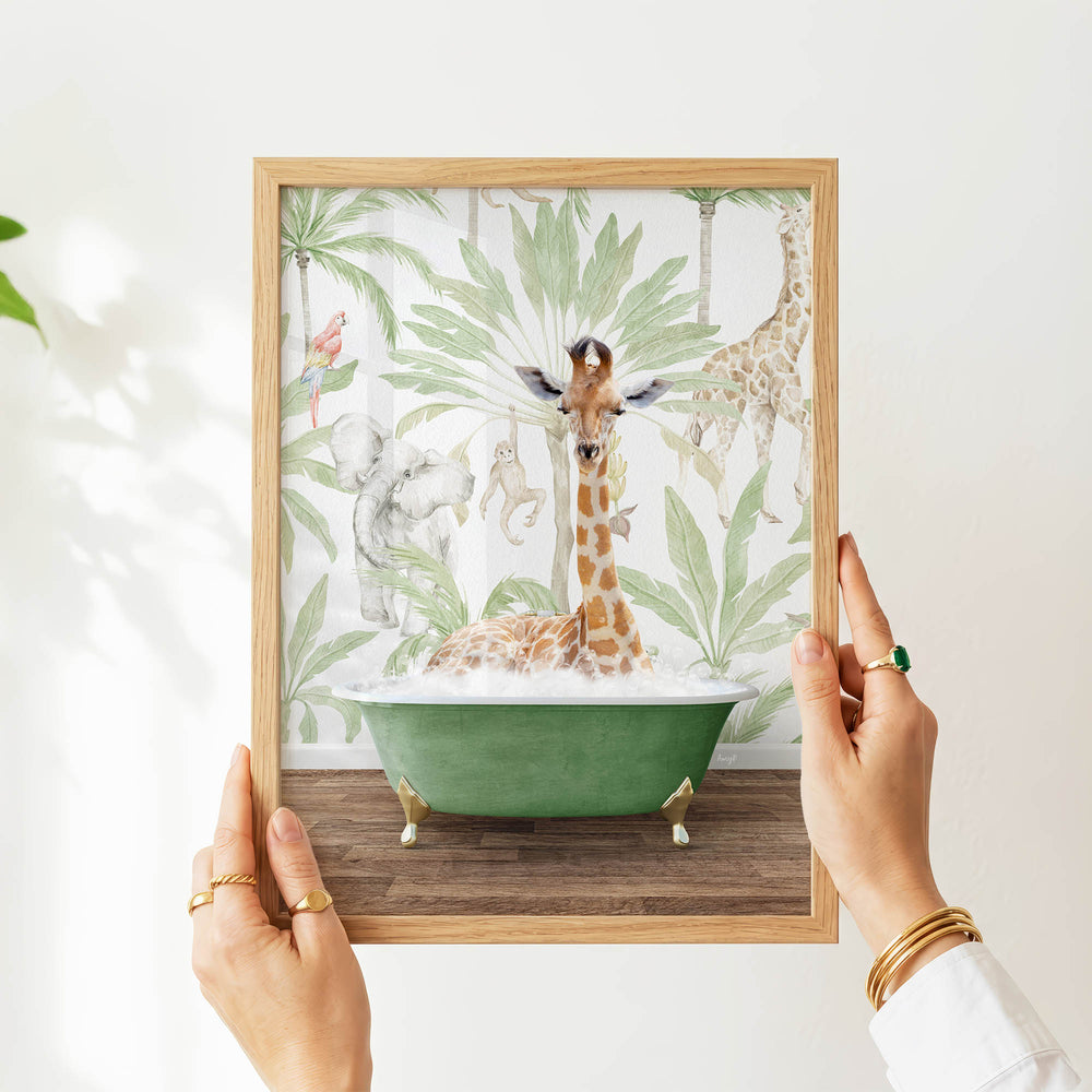 A person's hands are holding a framed picture of a giraffe sitting in a green bathtub, surrounded by tropical foliage.