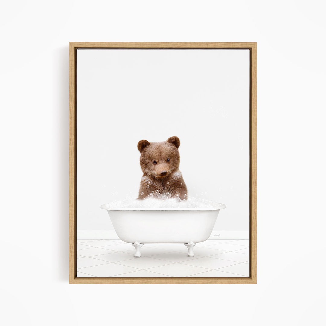A cute brown bear sitting in a white bathtub filled with bubbles, looking directly at the camera.