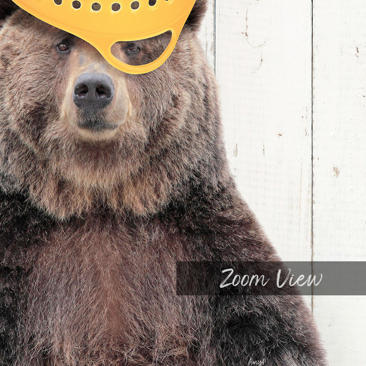 A close-up of a brown bear wearing a yellow plastic hat, with a wooden background.