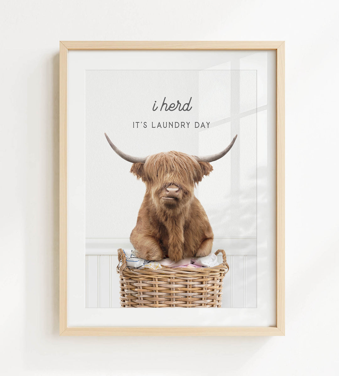 A framed image of a Highland cow sitting in a wicker basket, with the text "It's laundry day" above it.