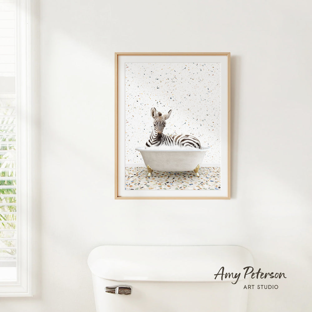 A framed print of a zebra sitting in a bathtub is hanging on a white wall above a white toilet.