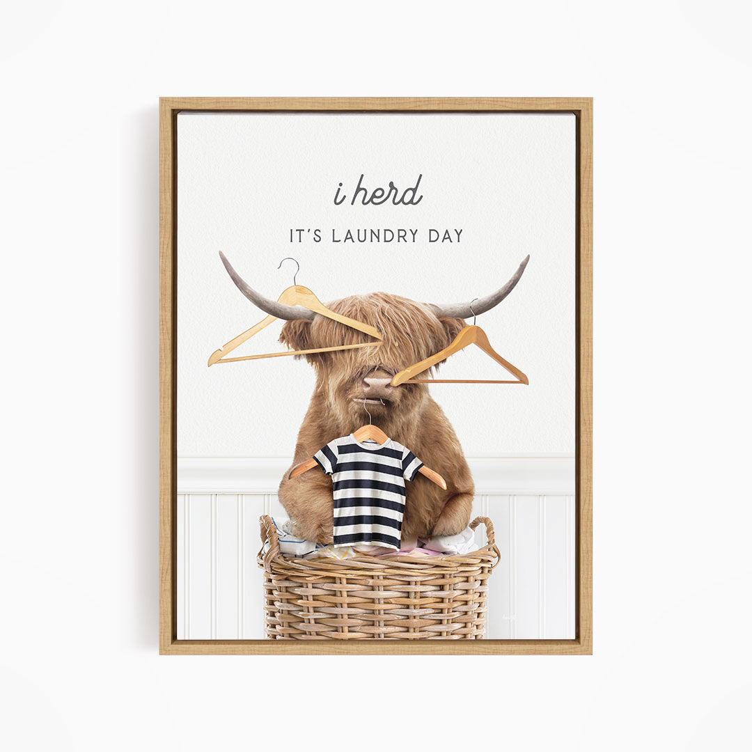 A framed image depicting a large, horned animal, possibly a yak or a yak dog, sitting in a wicker laundry basket and holding a pair of clothes hangers. The text above the animal reads "i herd, it's laundry day".