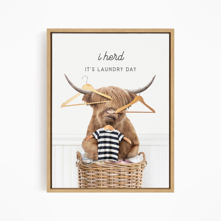 A framed image depicting a large, horned animal, possibly a yak or a yak dog, sitting in a wicker laundry basket and holding a pair of clothes hangers. The text above the animal reads "i herd, it's laundry day".