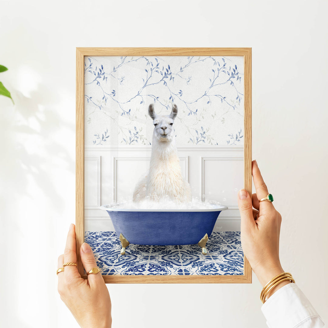 A person's hands are holding a framed picture of a white llama sitting in a blue bathtub filled with bubbles, against a white wall with a blue floral pattern.