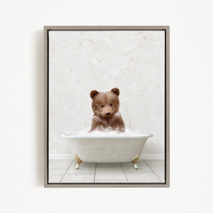 A cute brown bear cub sitting in a white bathtub filled with bubbles, looking directly at the camera.