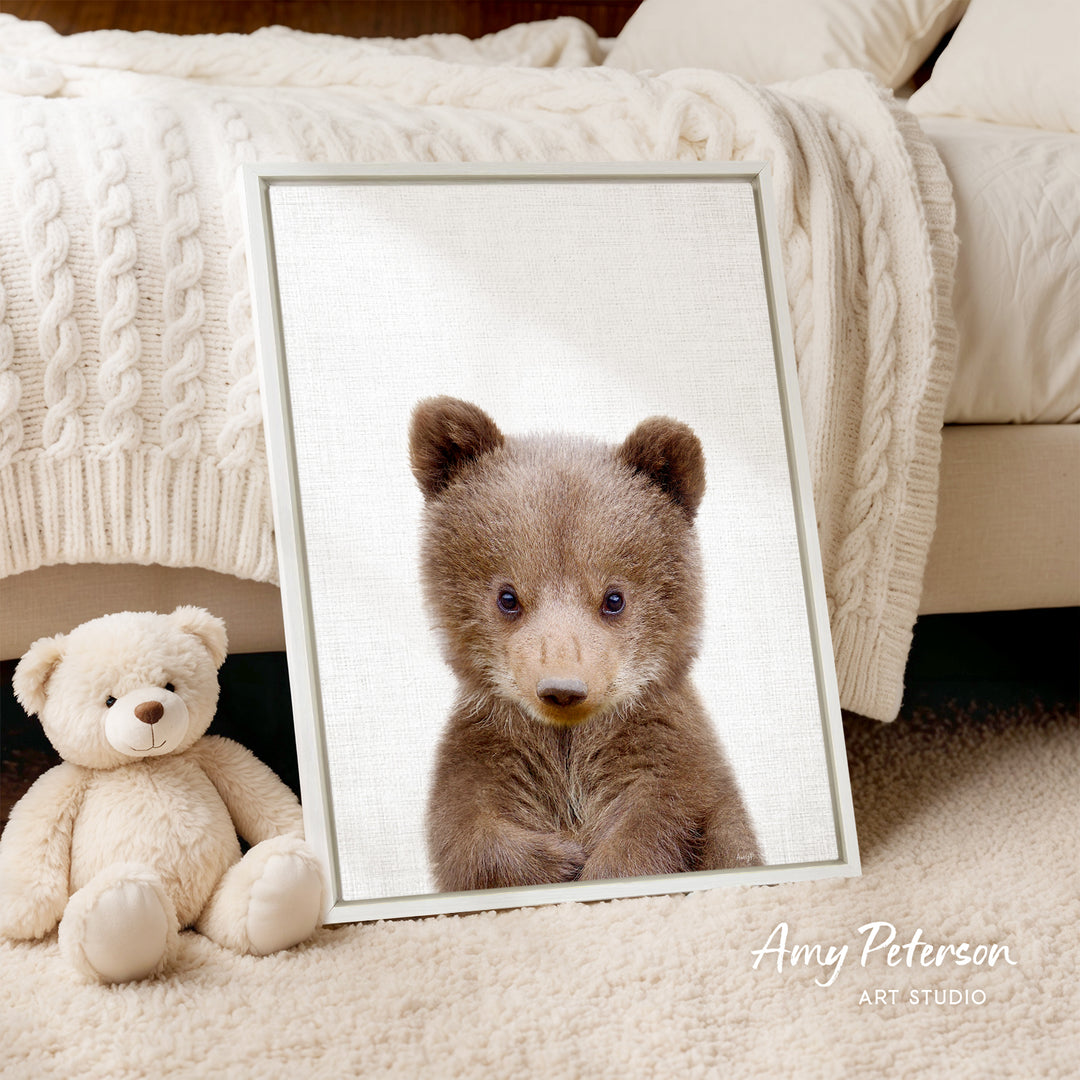 A framed image of a brown bear is displayed on a white bedspread, with a white teddy bear sitting next to it.