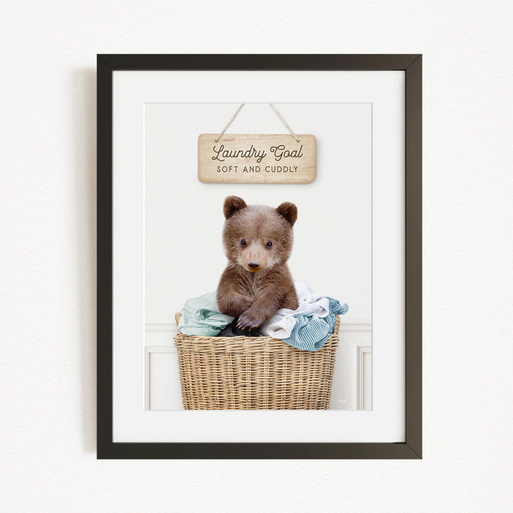 A framed image of a baby bear sitting in a wicker laundry basket, with a wooden sign above it that says "Laundry Goal Soft and Cuddly".