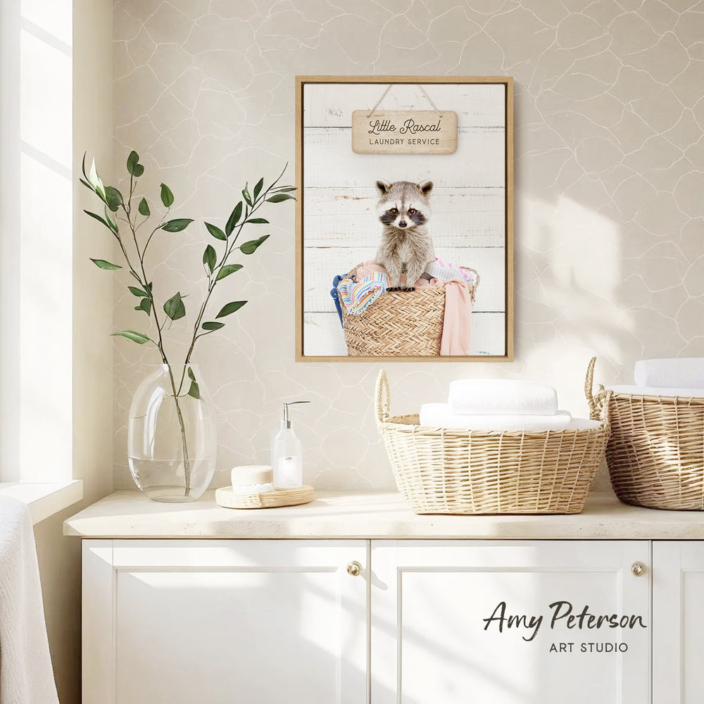 a framed picture of a raccoon sitting in a laundry basket, with a sign above it that reads "Little raccoon laundry service". The scene is set in a bathroom with a white cabinet and a vase of greenery.