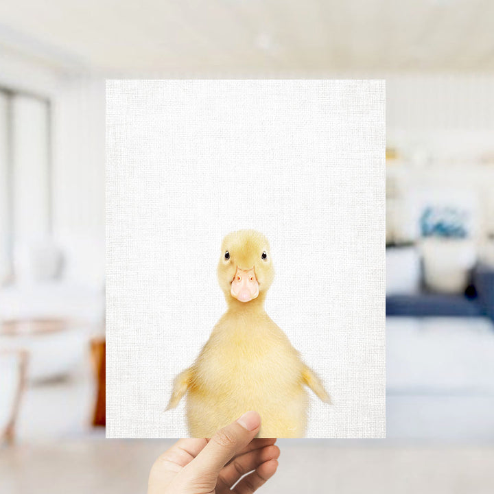 A hand is holding up a white square with a cute yellow duck on it, against a blurred background of a living room.
