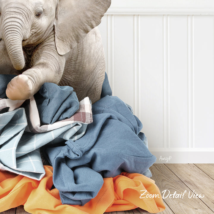 A baby elephant sitting on a pile of clothes, with a white wall and wooden floor in the background.