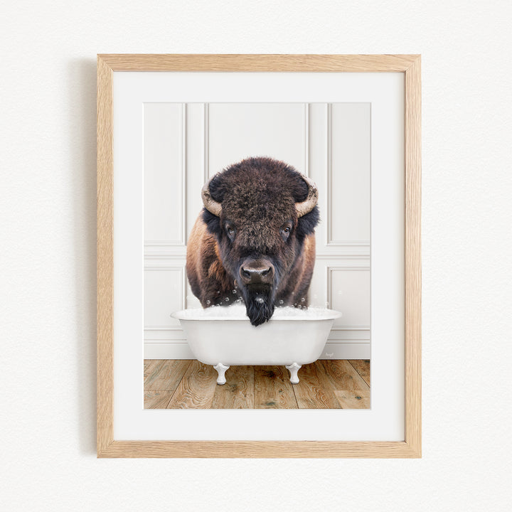 A buffalo with a black mane and nose is sitting in a white bathtub, with its head poking out of the tub.