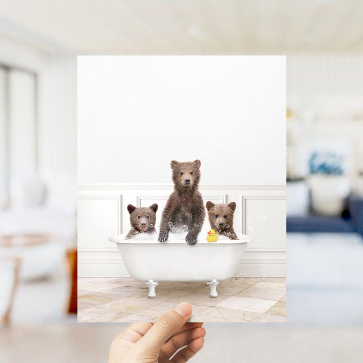 A hand holds up a framed photograph of three adorable brown bears sitting in a white bathtub, with one bear holding a yellow rubber duck.