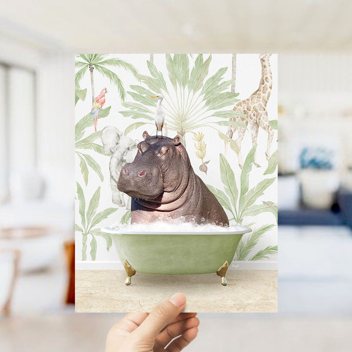 A hand holds up a framed image depicting a hippopotamus sitting in a bathtub surrounded by tropical foliage, including a giraffe and palm trees.