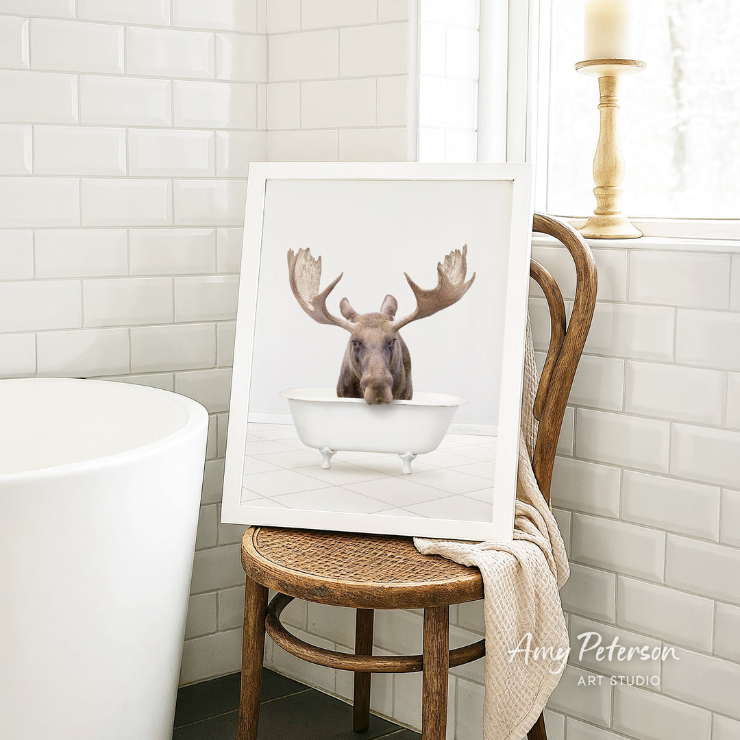 A framed image of a moose sitting in a bathtub is displayed on a wooden chair in a bathroom setting.