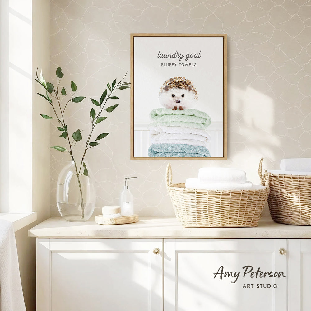 a framed poster of a hedgehog on a shelf, with a basket of towels below it. The poster has the text "laundry goal" and "fluffy towels" written on it. The shelf also has a vase with leaves and a bottle of soap.