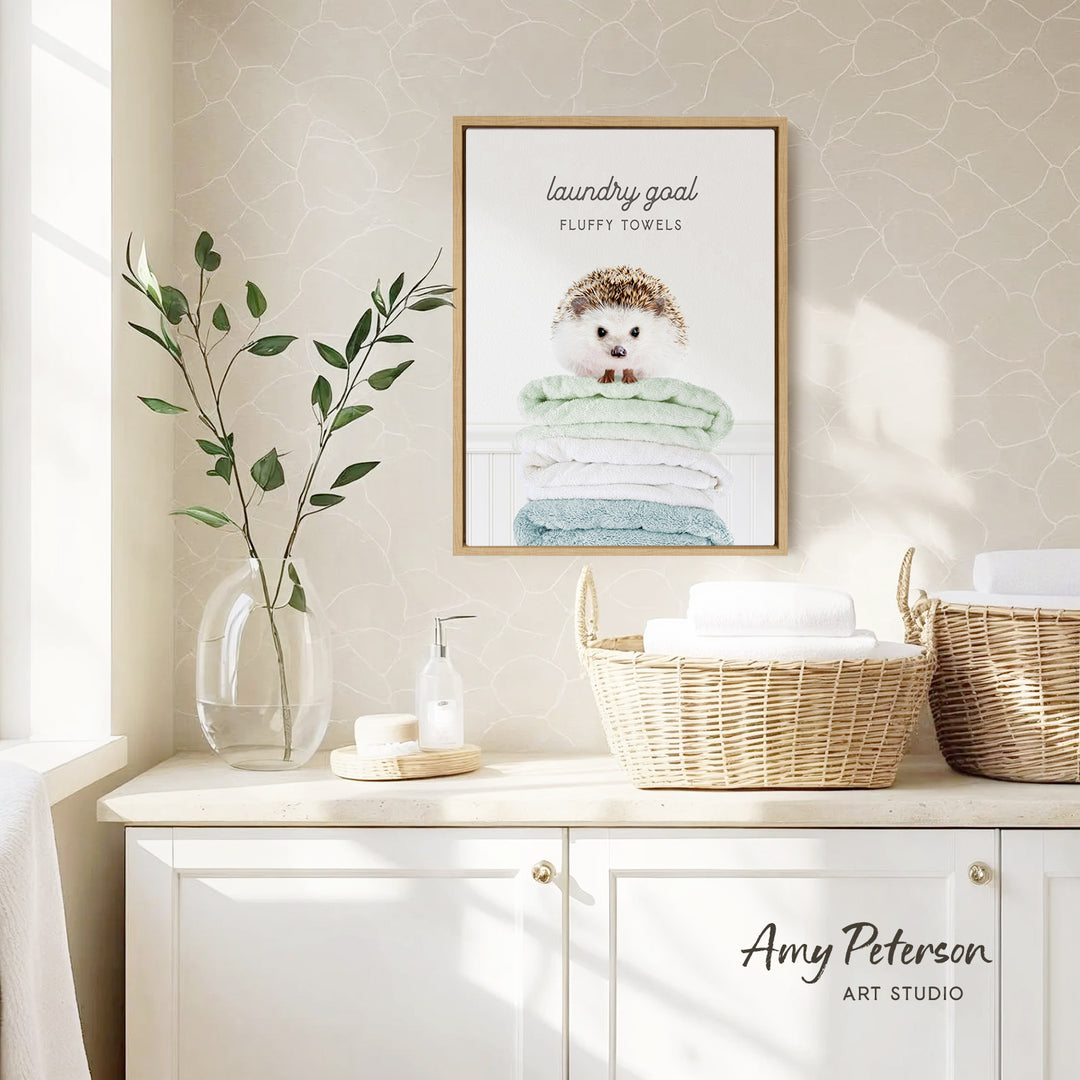 a framed poster of a hedgehog on a shelf, with a basket of towels below it. The poster has the text "laundry goal" and "fluffy towels" written on it. The shelf also has a vase with leaves and a bottle of soap.